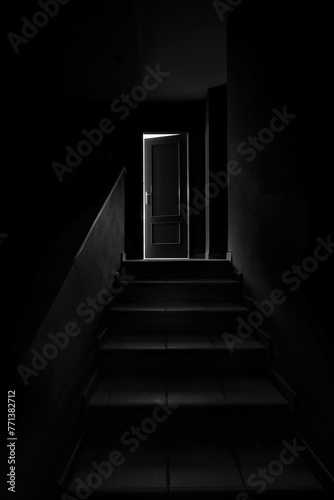 Black and white image of a dark staircase with a door ajar at the end, like a scene from a horror movie