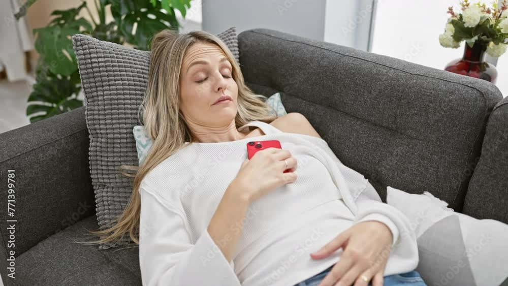 A young blonde woman checks her phone upon waking up in her modern living room. A young blonde woman checks her phone upon waking up in her modern living room.