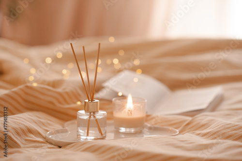 Liquid home perfume in glass bottle with bamboo sticks and open paper book with scented candle on ceramic tray in bed over glowing lights. Cozy atmosphere. © morrowlight