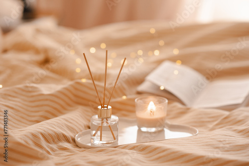 Liquid home perfume in glass bottle and scented candle with bamboo sticks and open paper book on ceramic tray in bed over glowing lights. Cozy atmosphere. © morrowlight