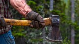 A close-up shot of a person's rough, gloved hands holding an axe, set against a natural forest background, depicting manual labor or lumber work.
