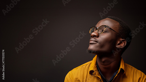 Handsome young African man wearing glasses and yellow shirt looking up isolated on dark background with copy space