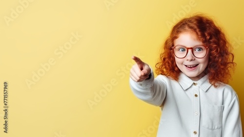 red-haired smiling schoolgirl with a backpack points her finger to the side on a yellow background, space for text