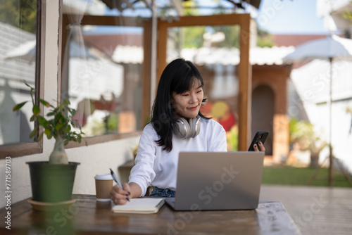 Wallpaper Mural Young woman wearing headphones holds a notebook and uses a laptop at an outdoor cafe table. Finding and thinking of ideas, writing them down in a notebook Torontodigital.ca