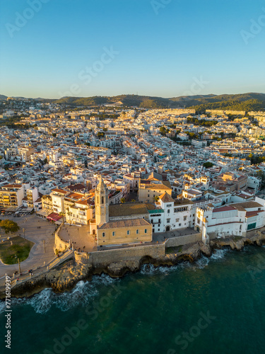Drone point of view of Sitges City at sunset. Panoramic view of all city. Waves splashing the rocks and buildings. Travel destination. Beautiful warm colours at sunset, reflecting on buildings.