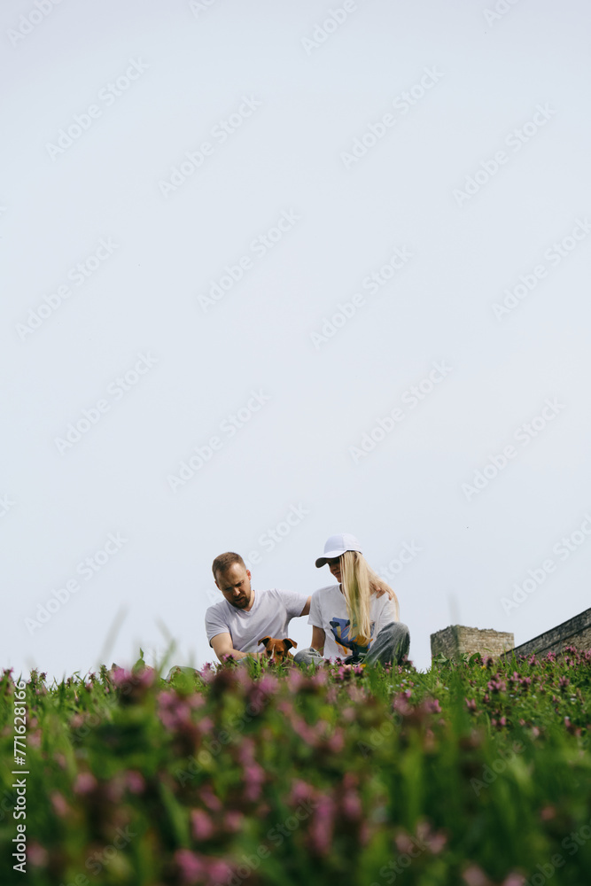 Young couple sitting in a green clearing among wildflowers and enjoying a peaceful walk with a Jack Russell terrier. People with a dog in Kalemegdan Park near the walls of the old historical fortress. Young couple sitting in a green clearing among wildflowers and enjoying a peaceful walk with a Jack Russell terrier. People with a dog in Kalemegdan Park near the walls of the old historical fortress.