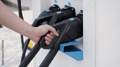 Hand holding EV charging plug in front of camera with blurred background of outdoor. Driver of electric car hold batteries charger plug. Sustainable energy powered electric charging station background