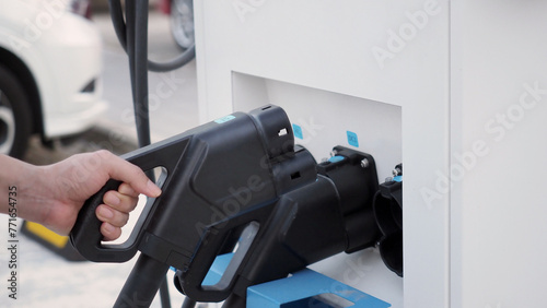 Hand holding EV charging plug in front of camera with blurred background of outdoor. Driver of electric car hold batteries charger plug. Sustainable energy powered electric charging station background