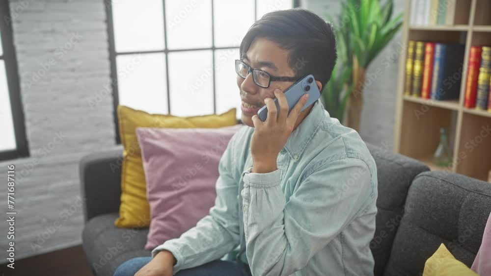 An asian man converses on a smartphone in a modern living room, portraying casual home life. An asian man converses on a smartphone in a modern living room, portraying casual home life.