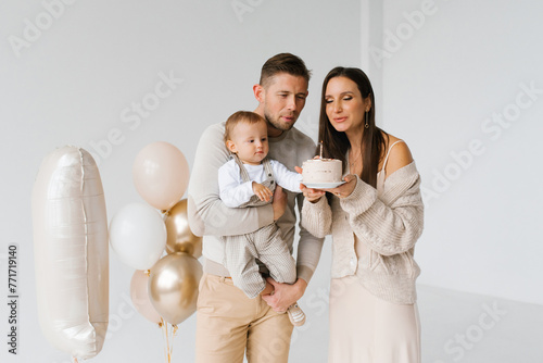 Young family with a one-year-old son blows out a candle on a birthday cake. The scene is joyful and festive