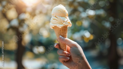 Hand holding a vanilla ice cream cone against a bokeh background of sunlight through trees. Summer treat and nature concept