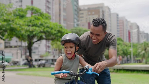 Happy latin father helping smiling boy to ride bike on cityu park. Happy latin child and young dad riding bike. Smiling daddy teaching son to ride a balance bicycle.
