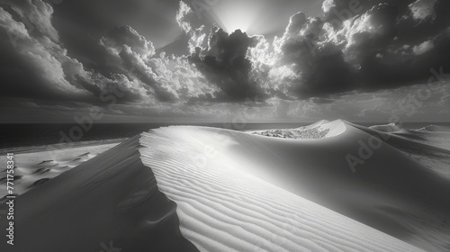  A monochrome image depicts a sand dune with sunlight filtered through cloudy skies