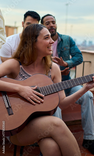 Vertical. Smiling Caucasian blonde woman happy plays flamenco guitar while her group multiracial friends clap at rooftop party. Young people gathered together enjoying summer vacations © CarlosBarquero