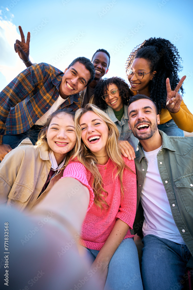 © CarlosBarquero - Vertical. Group of multiethnic friends taking selfie with smart mobile phone outdoors. Happy young multiracial people smiling together looking camera. University students having fun in college campus