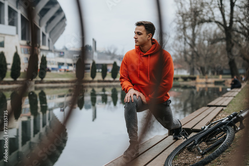 Contemplative young man resting next to his bicycle in a serene park setting with cloudy skies and a tranquil pond.