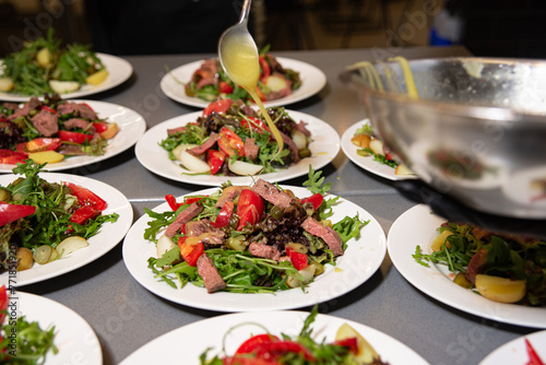 Chef Dressing Steak Salad Plates in Commercial Kitchen. Chef precisely drizzling dressing over steak salads, beautifully arranged on plates in a busy commercial kitchen setting.