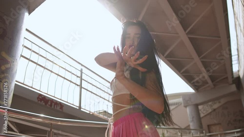 Slow motion. Low Angle Shot. A girl with long hair looks at the camera and moves her arms elegantly, while street dancing, while standing in front of the sun under a city pedestrian bridge outdoors photo