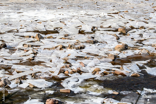 A shallow river covered with shards of broken ice supported by mud and rock. Some water flows among the ice.