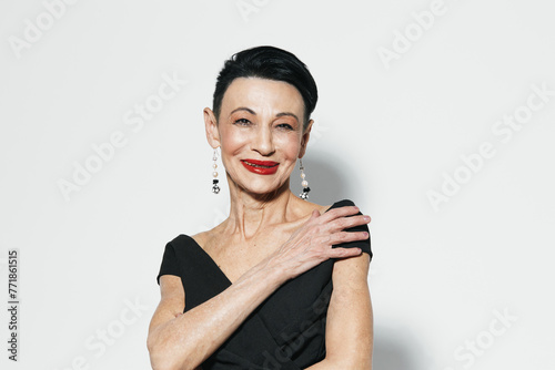 Elegant older woman in black dress and red lipstick posing in front of white wall