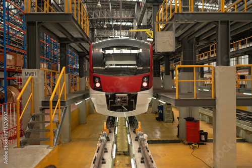 Front view of a red and white modern train situated on tracks in a maintenance depot, with industrial equipment around.