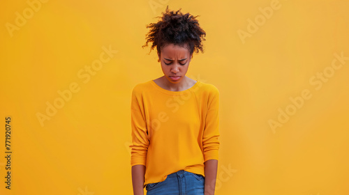 A young African American woman stands with her head down, looking sad and dissapointed against an isolated yellow background photo