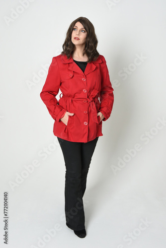 full length portrait of beautiful brunette woman model, wearing red trench coat jacket leather pants. standing pose, walking towards the camera. isolated on white studio background.