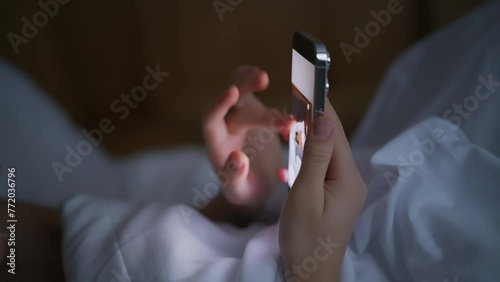 The girl is on her cell phone. A girl flipping through photos on her smartphone. A young girl is lying in bed and looking at pictures on her phone. Cell phone. Processing photos on the phone. White