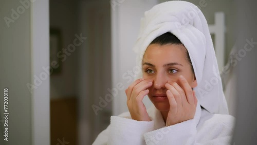 The girl is getting a facial massage. Girl in front of mirror giving massage under eyes. Gentle facial massage. Self-care. Massage with fingers of hands. Bathroom. White robe. Hotel room. Morning