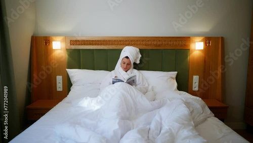 a girl reading a book. Girl after showering read a book in bed in her bathrobe. Hotel room. Bright cozy room. White bed. Morning. A girl resting in bed and reading a magazine. A young girl with a