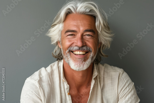 A man with white hair and a white shirt is smiling. He is sitting on a grey surface. Attractive 50 year old smiling man with medium white hair in cream-colored clothes against a gray background. © Nataliia_Trushchenko