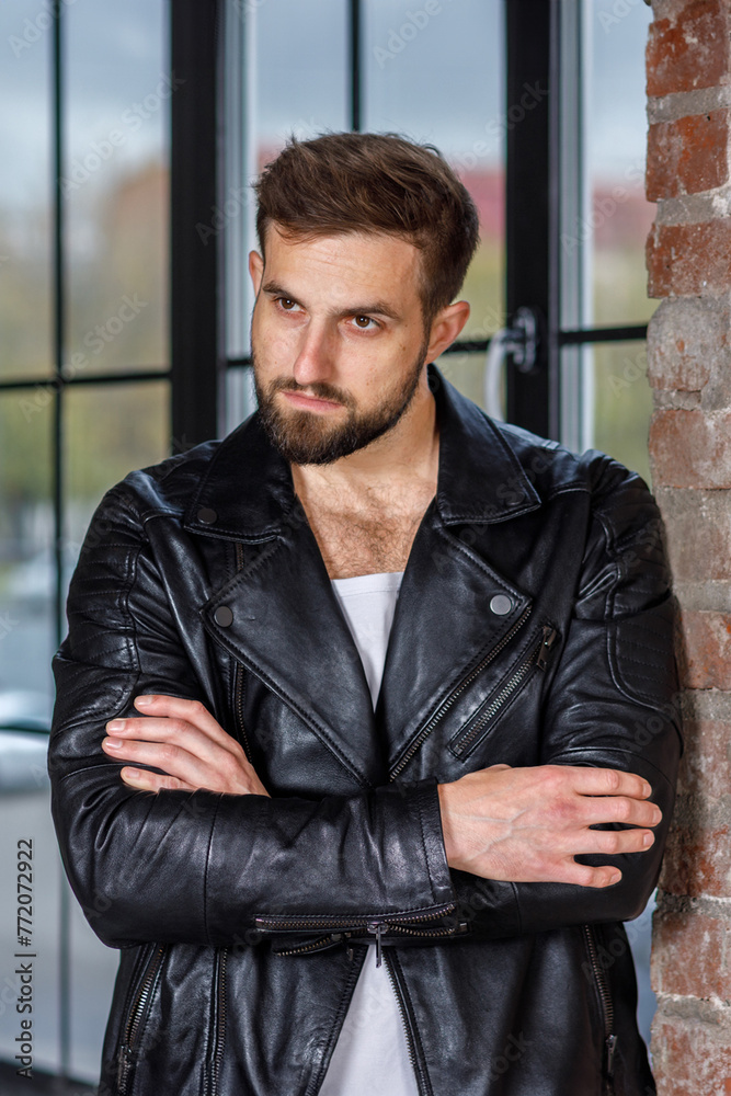 Obraz premium Modern young man with a stylish hairstyle, wearing a cool black leather jacket and standing on a window