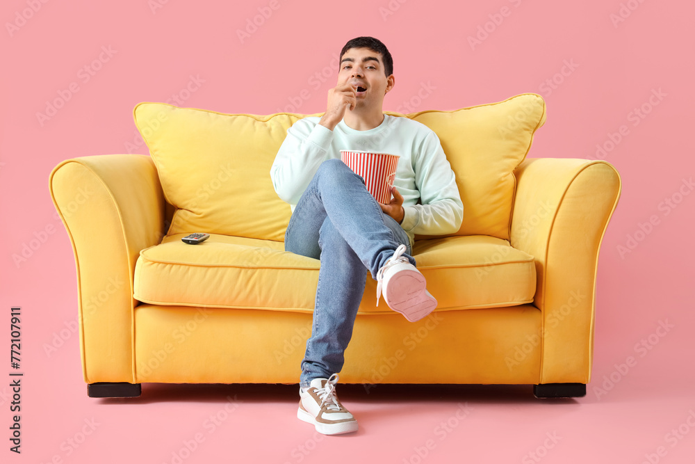 Handsome young man with popcorn watching TV on yellow sofa against pink background Handsome young man with popcorn watching TV on yellow sofa against pink background