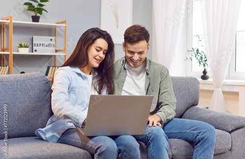 Young happy couple using laptop sofa sitting, enjoy watching movie, streaming service, shopping online. Small family at home choose product via internet to order online, friendly virtual call meeting
