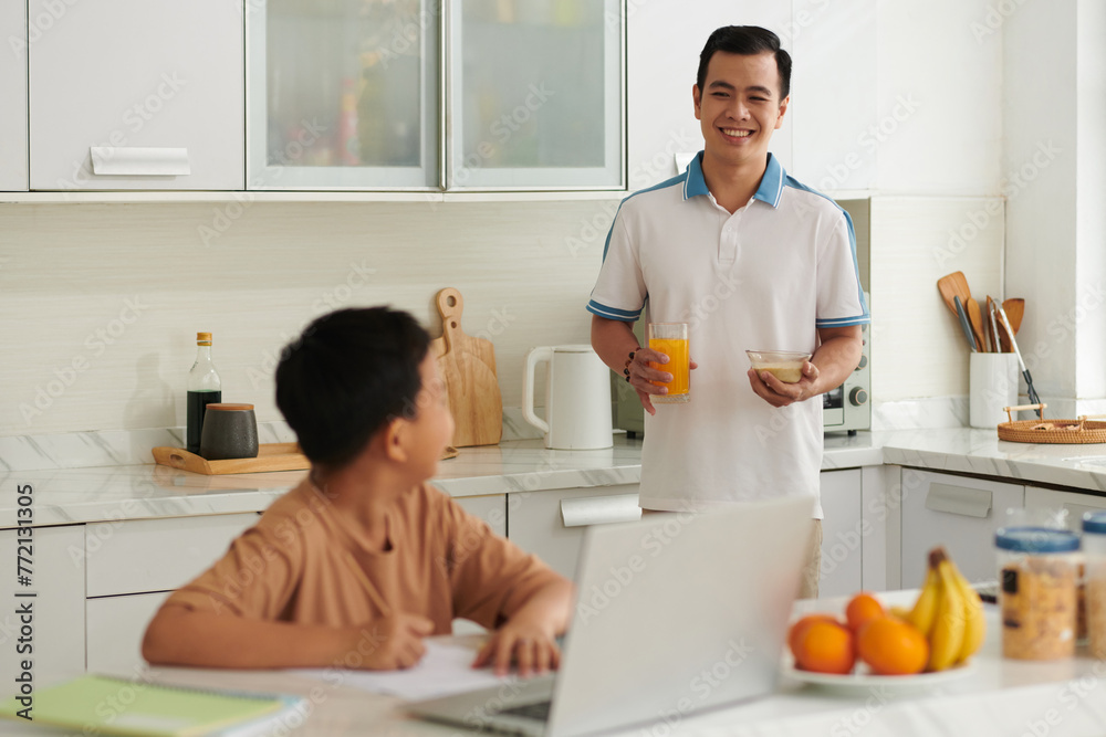Smiling father bringing glass of juice and porridge to son doing homework at kitchen counter Smiling father bringing glass of juice and porridge to son doing homework at kitchen counter