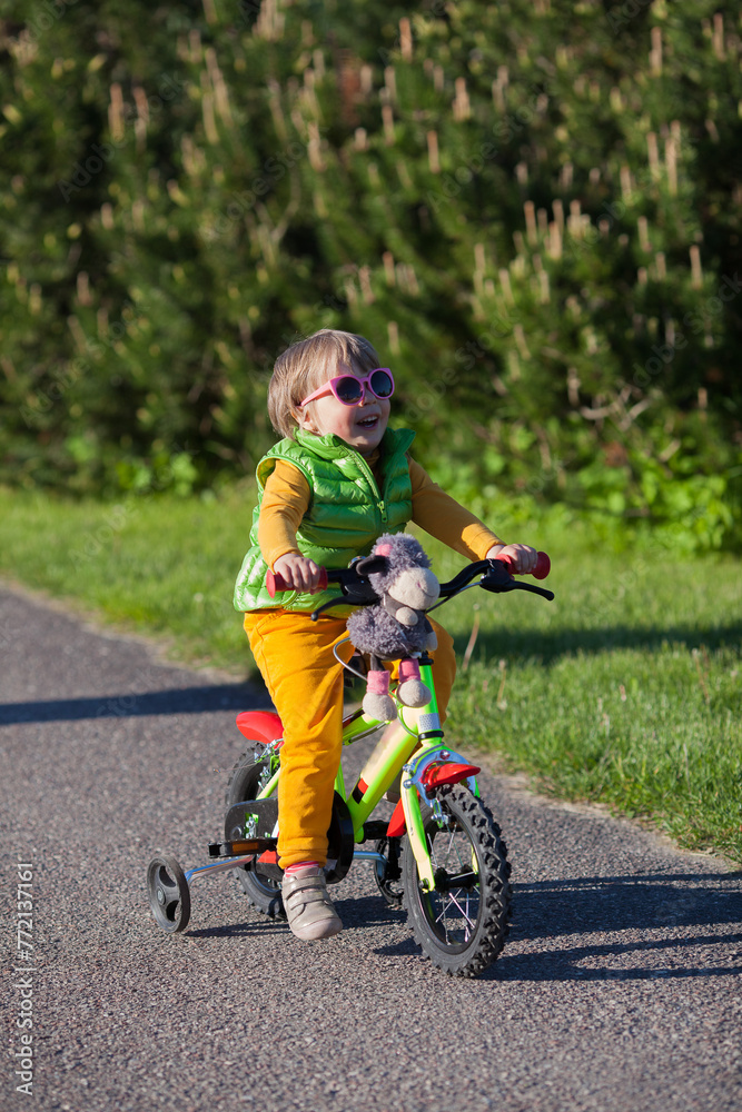 A little cute girl is riding on a bike with her soft toy in summer. Green and orange kid's outfit. Vertical image A little cute girl is riding on a bike with her soft toy in summer. Green and orange kid's outfit. Vertical image
