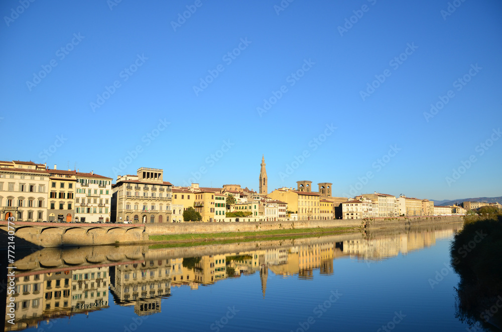 A distant view of traditional Italian houses by the riverbank of Arno River in Pisa against a cloudless sky. The background texture features village homes in Europe with water reflection