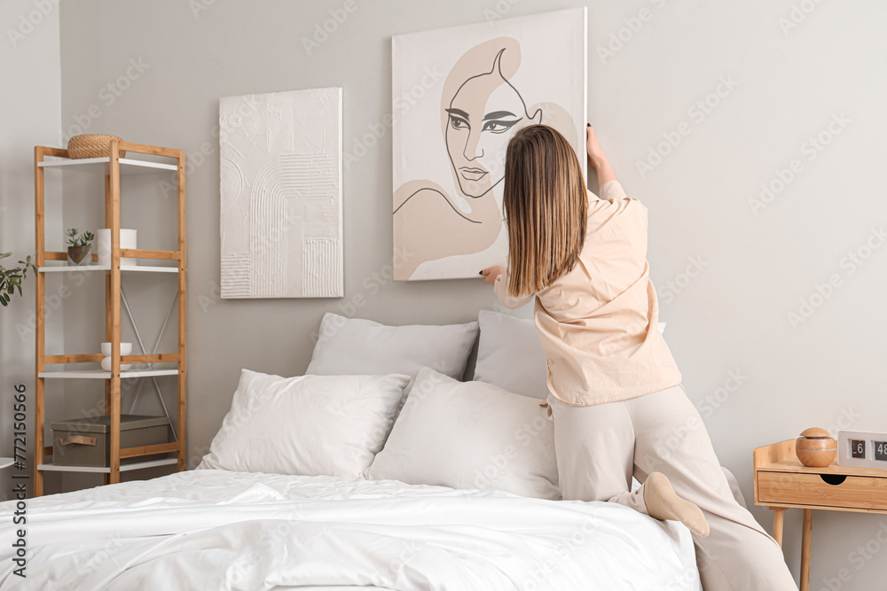 Young woman hanging stylish painting on light wall in bedroom, back view Young woman hanging stylish painting on light wall in bedroom, back view