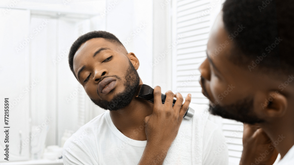 Man trimming beard with electric clippers in mirror in bathroom - obrazy, fototapety, plakaty 