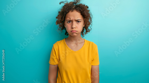 Sad teenage girl standing isolated on blue background, frowning and looking at camera with sad expression in yellow tshirt, school kid crying