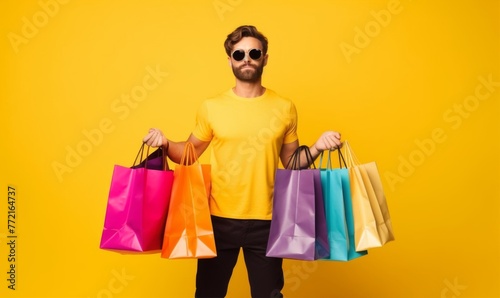 a guy in black pants and a yellow T-shirt walks with colorful bags in his hands, adjusts his sunglasses on a white background 