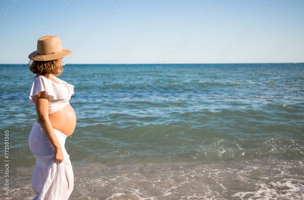 Pregnant woman with sun hat during her walk on the beach relaxing on her summer vacation, happy pregnancy outdoors.