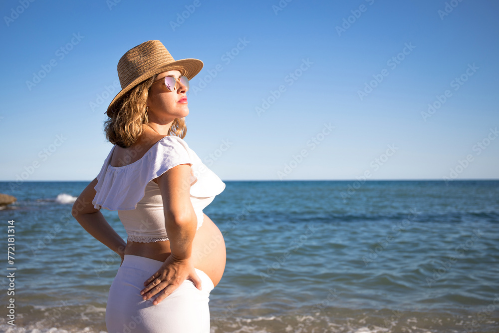 Pregnant woman with sun hat during her walk on the beach relaxing on her summer vacation, happy pregnancy outdoors.
