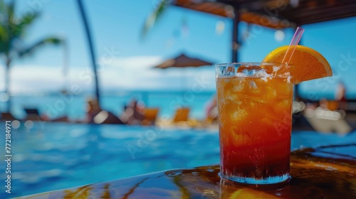A glass of orange juice with a straw in it sits on a table by a pool. The drink is garnished with an orange slice. The scene is set in a beach or poolside setting, with people enjoying the sun