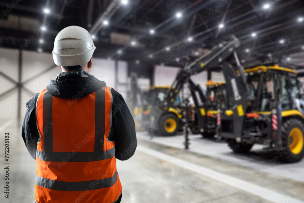Obraz premium Builder in hangar with construction equipment. Man working with his back to camera. Machines for construction and engineering work. Construction worker in reflective vest. Builder chooses bulldozer