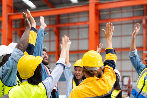 group of diverse workers raising hands to celebrate project success at manufacturing industrial factory, team colleagues in safety clothes smile glad shouting about production progress in the meeting