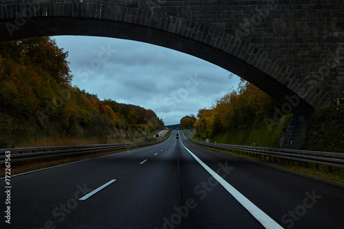 The arch of the bridge over the road stretching into the distance. Trees and shrubs with autumn foliage on the side of the road. Germany.