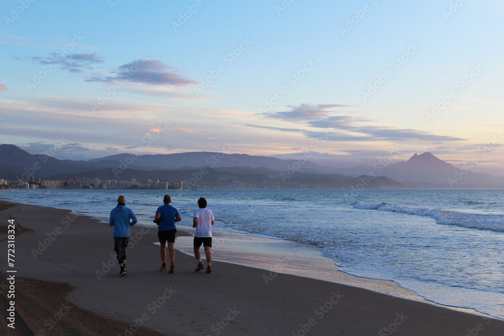 three men jogging on the beach against the background of the sunrise over the sea