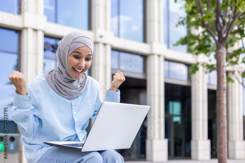 A joyful Muslim woman in a hijab raises her fists in success while using a laptop outdoors near modern office buildings.