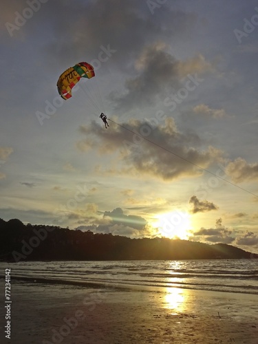 The tranquil ocean at sunset, framed by a dark mountain silhouette, gentle light peeking through the clouds, and a paragliding adventure in the distance.